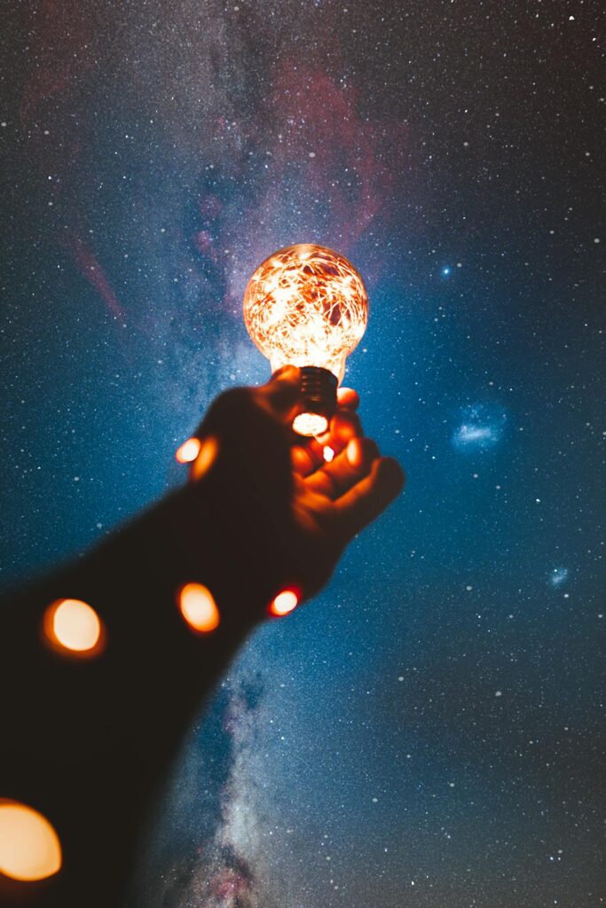 A hand holding an illuminated light bulb extends upward toward a starry night sky.