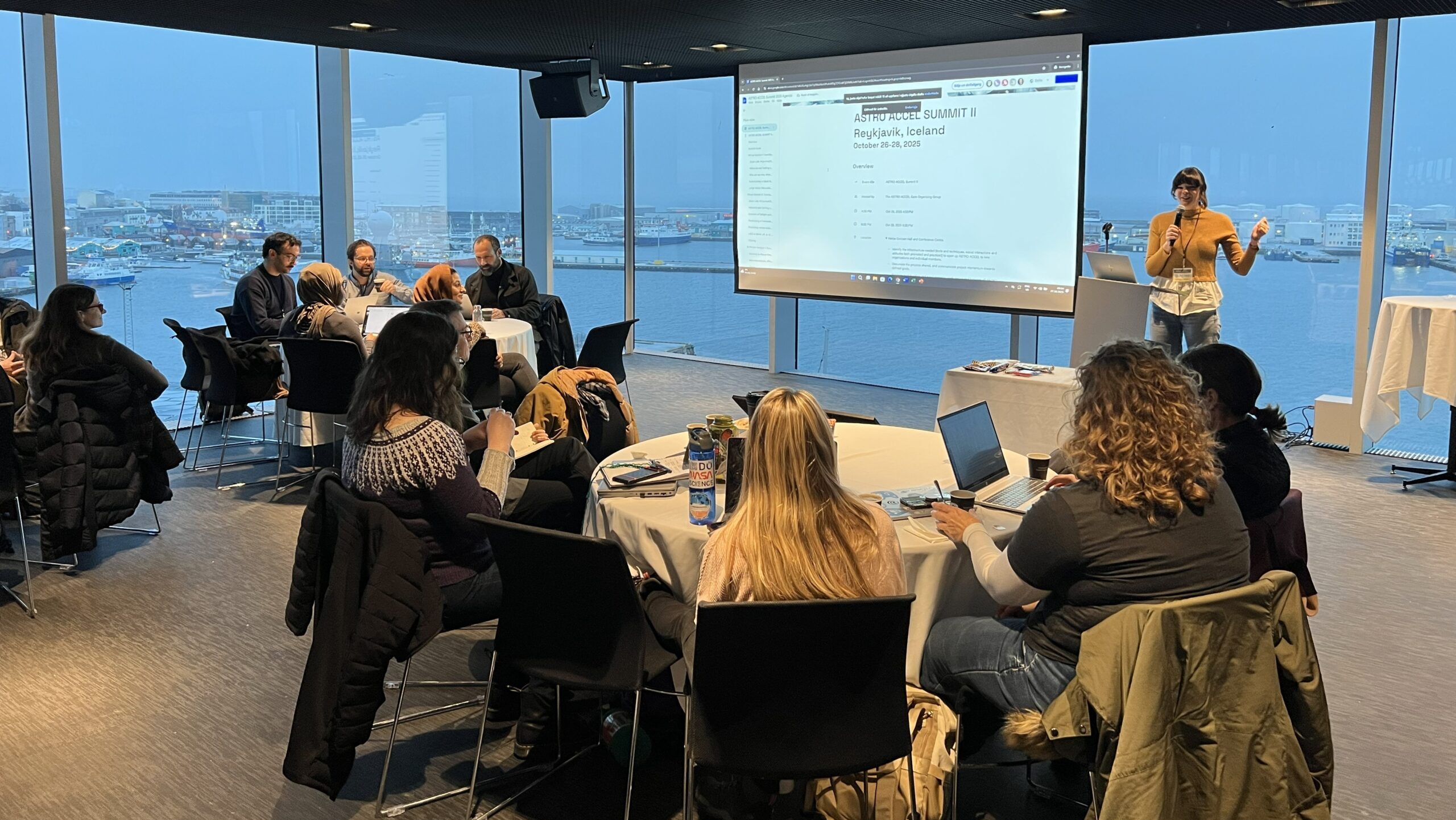 A presentation taking place in a modern conference room with a large window overlooking a harbor. Attendees are seated around tables with laptops, facing a presenter near a screen displaying a PowerPoint slide.