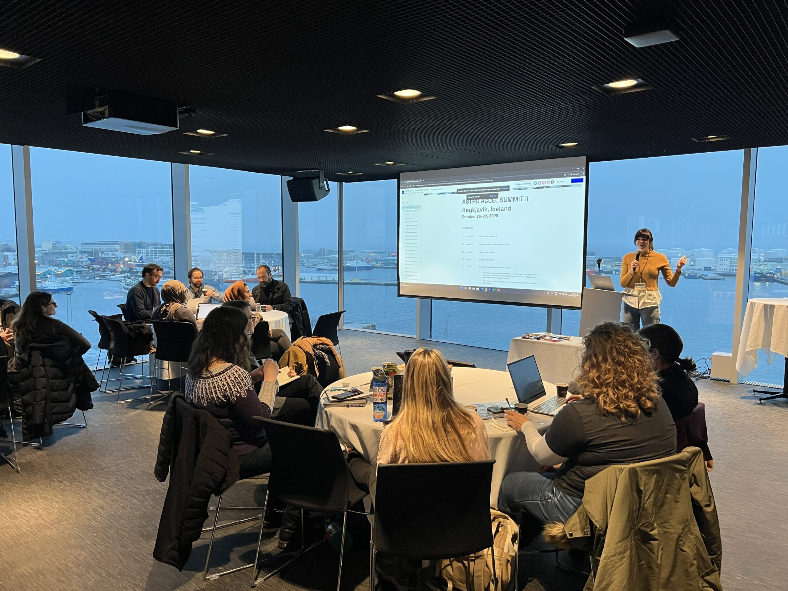 A presentation taking place in a modern conference room with a large window overlooking a harbor. Attendees are seated around tables with laptops, facing a presenter near a screen displaying a PowerPoint slide.