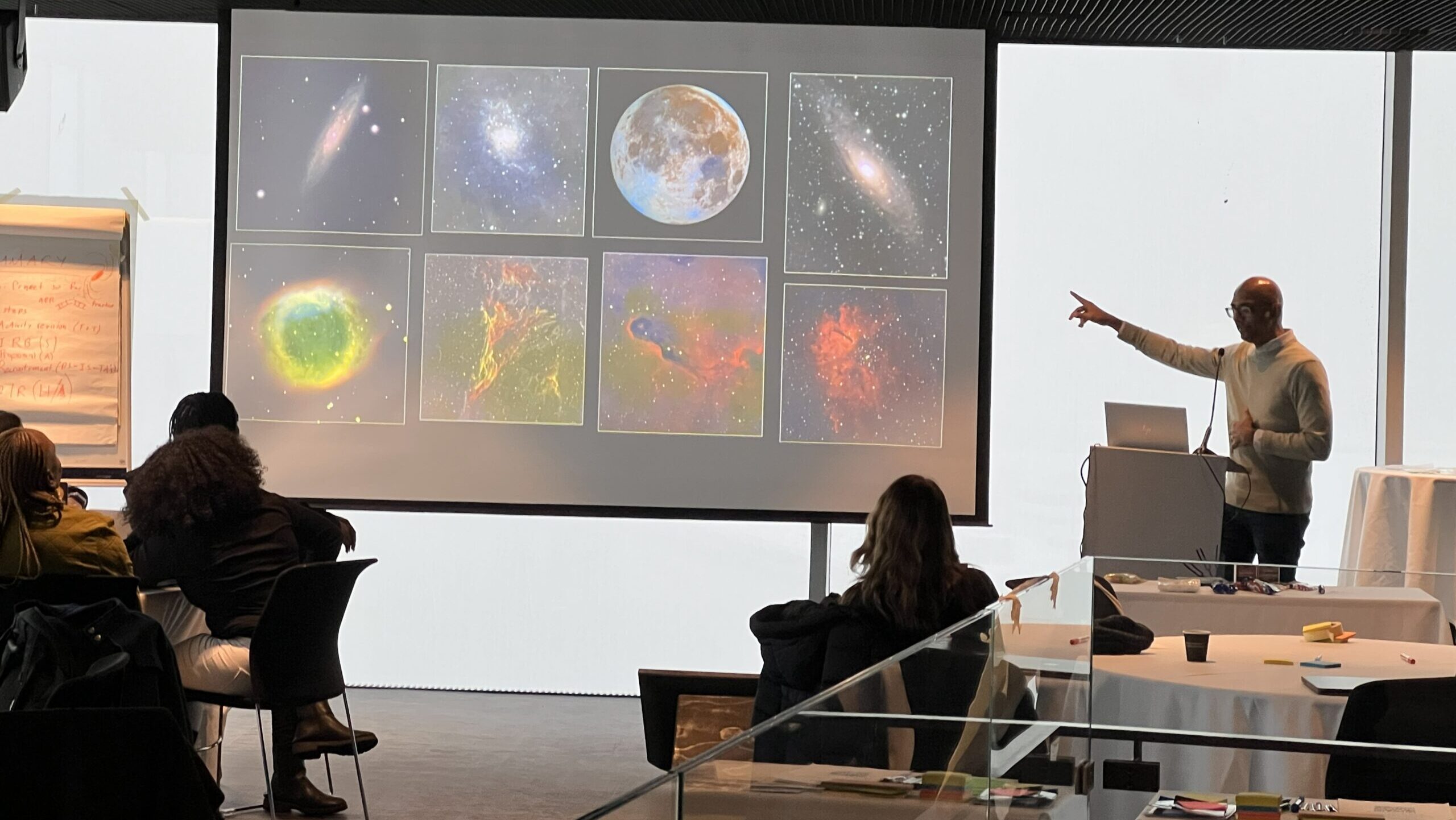 A presenter pointing at a large screen displaying various astronomical images during a lecture in a modern conference room with attendees seated at tables.