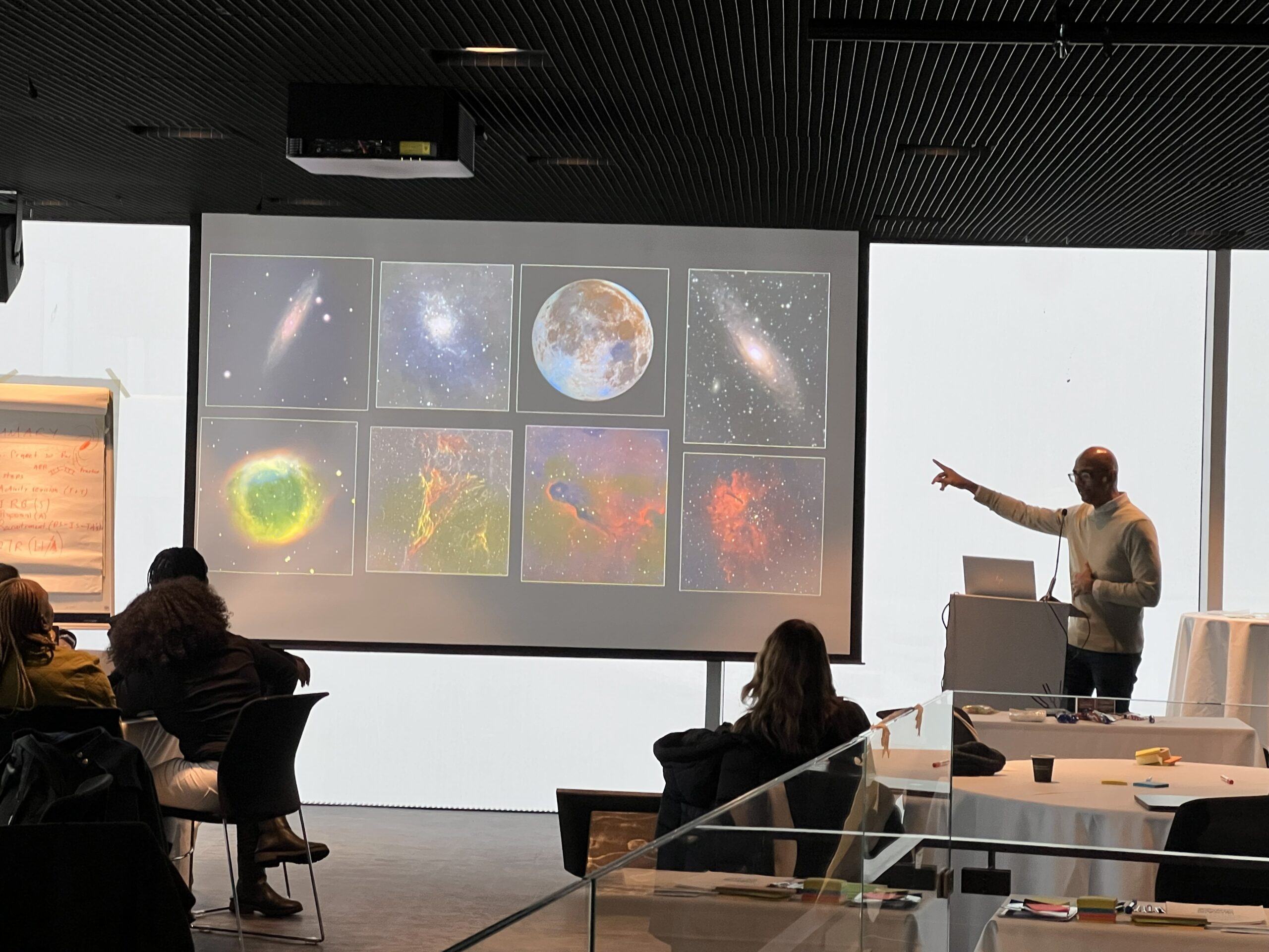 A presenter pointing at a large screen displaying various astronomical images during a lecture in a modern conference room with attendees seated at tables.
