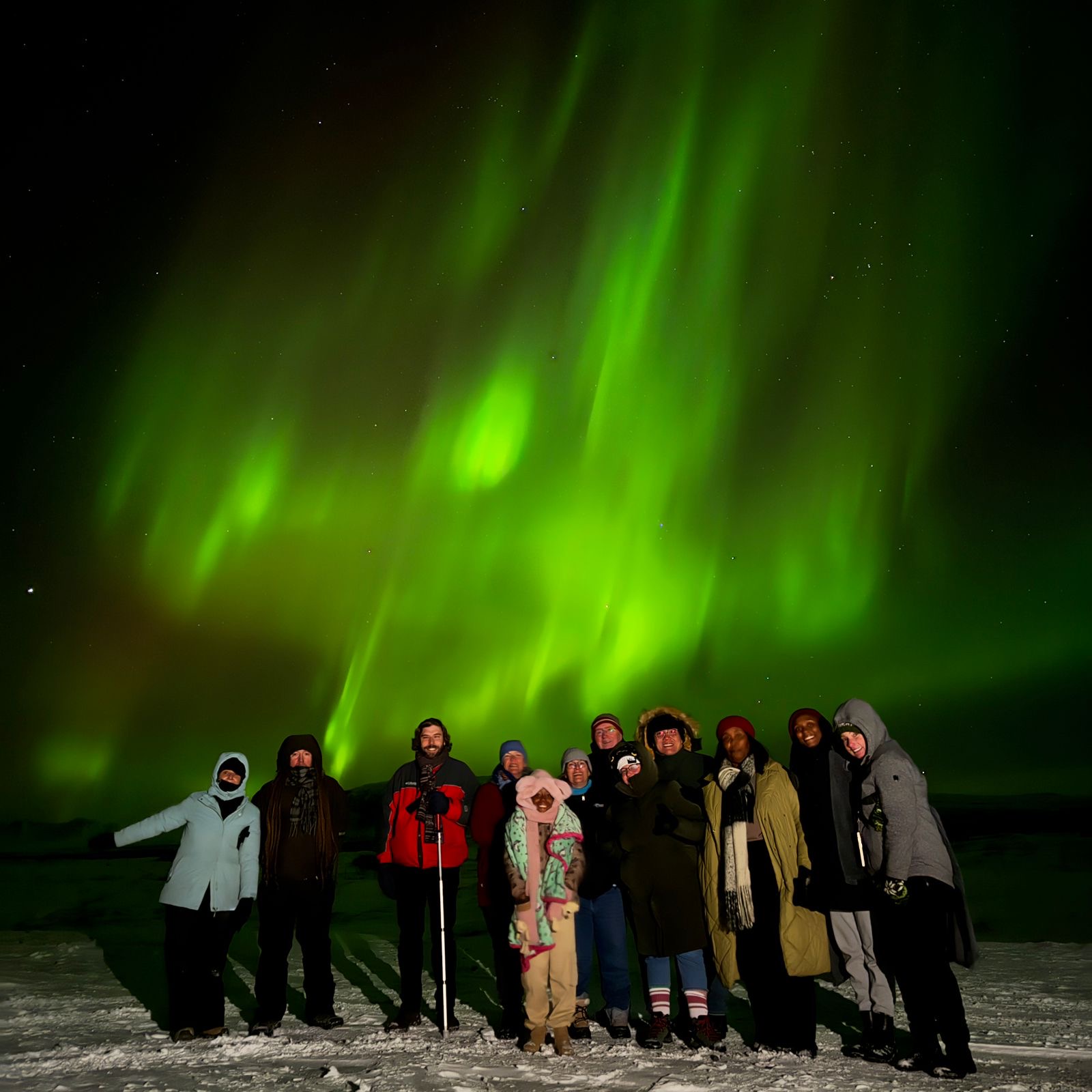 Group of ASTRO ACCEL members standing under the Northern Lights at night, posing for a photo on a snowy landscape.